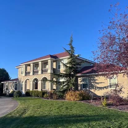 Elegant two-story house with a red tile roof, green lawn, and tall trees against a blue sky.